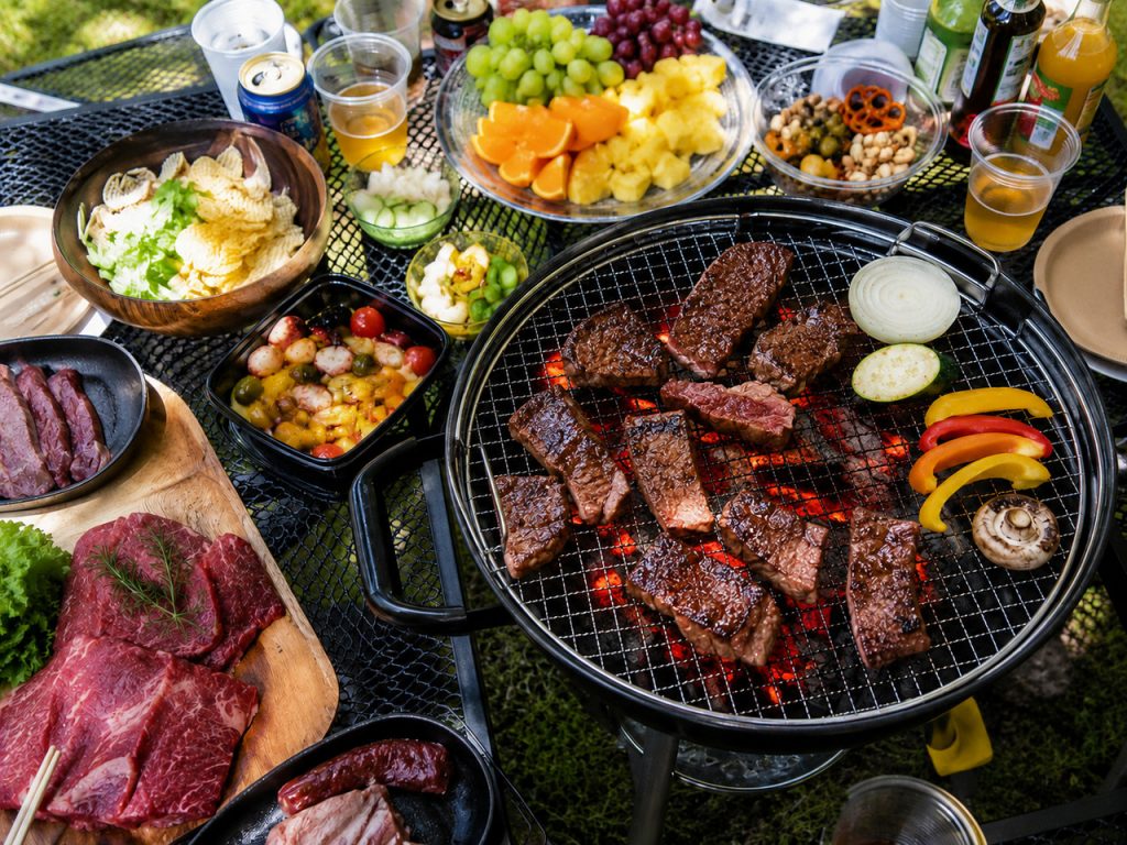 肉や野菜を焼いているバーベキューグリル、木のプレートに盛られた肉、フルーツ、アヒージョ、サラダなどがテーブルに並べられている