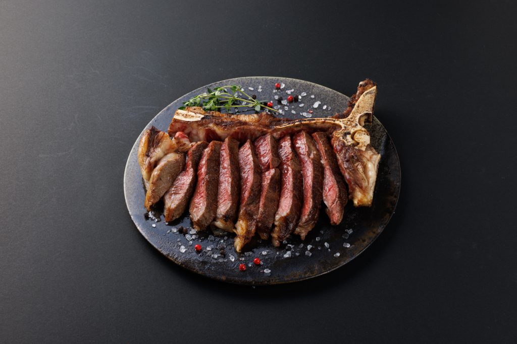 黒い皿に骨付きのステーキ肉が盛られていて、まわりに岩塩とピンクペッパーがのっている
